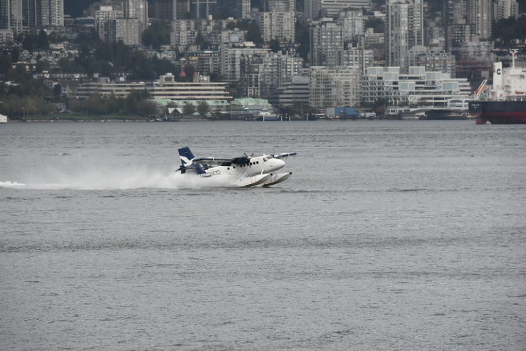 L'image montre un hydravion de la compagnie Harbour Air décollant ou atterrissant sur une étendue d'eau, probablement un port ou une baie. L'avion produit des éclaboussures derrière lui, indiquant qu'il est en mouvement rapide sur la surface de l'eau. En arrière-plan, on peut voir une ville avec de nombreux immeubles de grande hauteur, ainsi qu'une zone portuaire avec des bateaux et des structures industrielles. Le paysage urbain dense suggère qu'il s'agit d'une grande ville côtière.